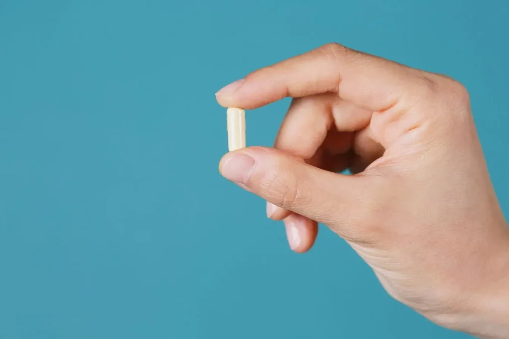 Une main tient délicatement une gélule jaune de vitamine B12 entre le pouce et l'index sur un fond bleu uni pour illustrer une supplémentation nutritionnelle.