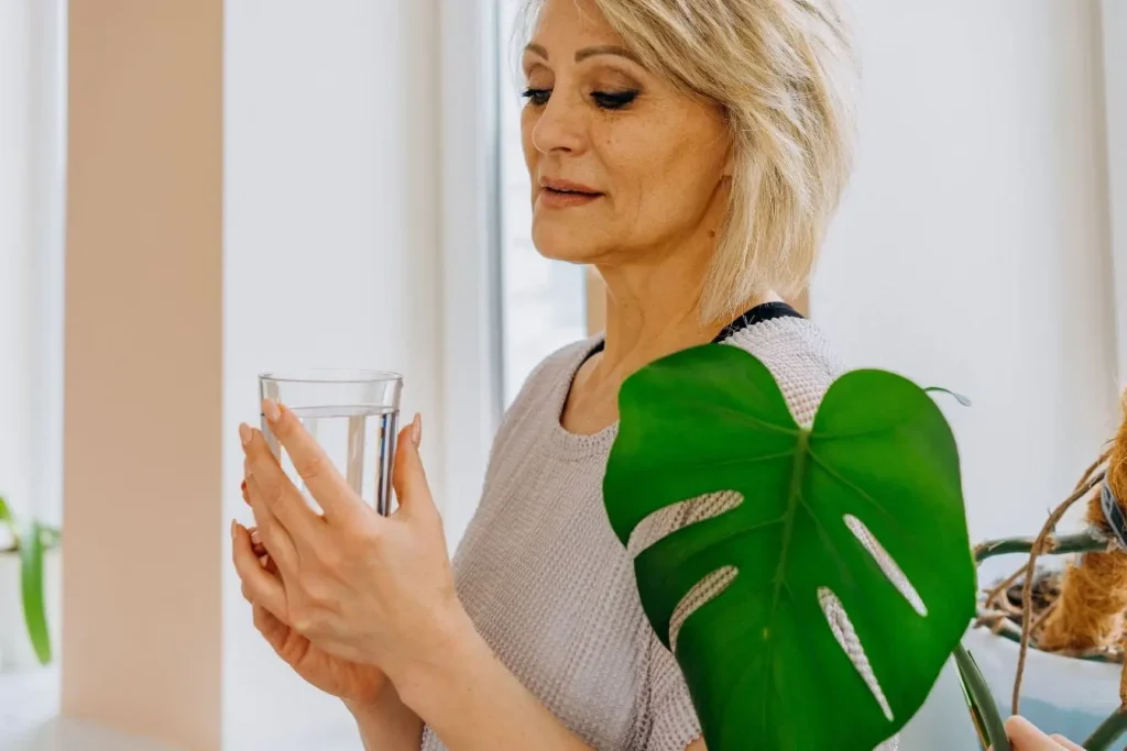 Femme tenant un verre d’eau près d’une plante verte, lumière douce, évoquant la recherche de bien-être face aux symptômes de la ménopause précoce.