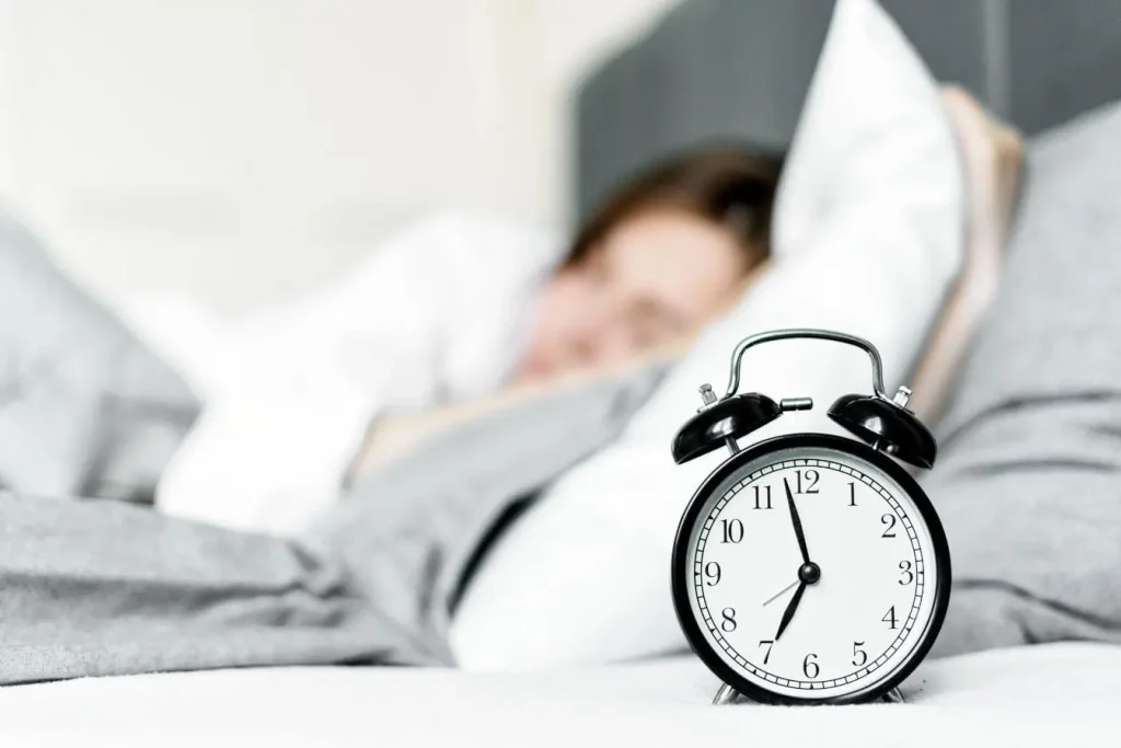 Réveil noir posé sur lit avec personne endormie, image qui illustre la fragilité du sommeil face aux pauses respiratoires de l’apnée.