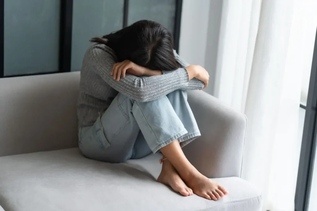 Personne assise sur canapé clair, genoux repliés et tête baissée, reflet d’un état de détresse lié au SSPT.
