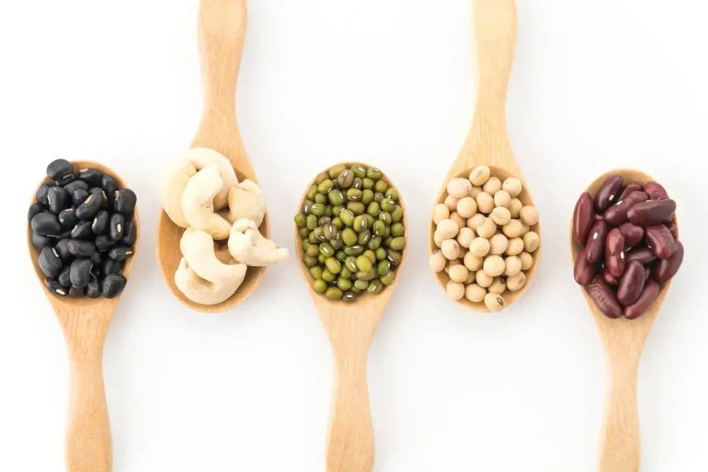 Cinq cuillères en bois alignées avec haricots noirs, noix de cajou, mungo verts, soja et haricots rouges, image qui illustre des aliments riches en tryptophane.