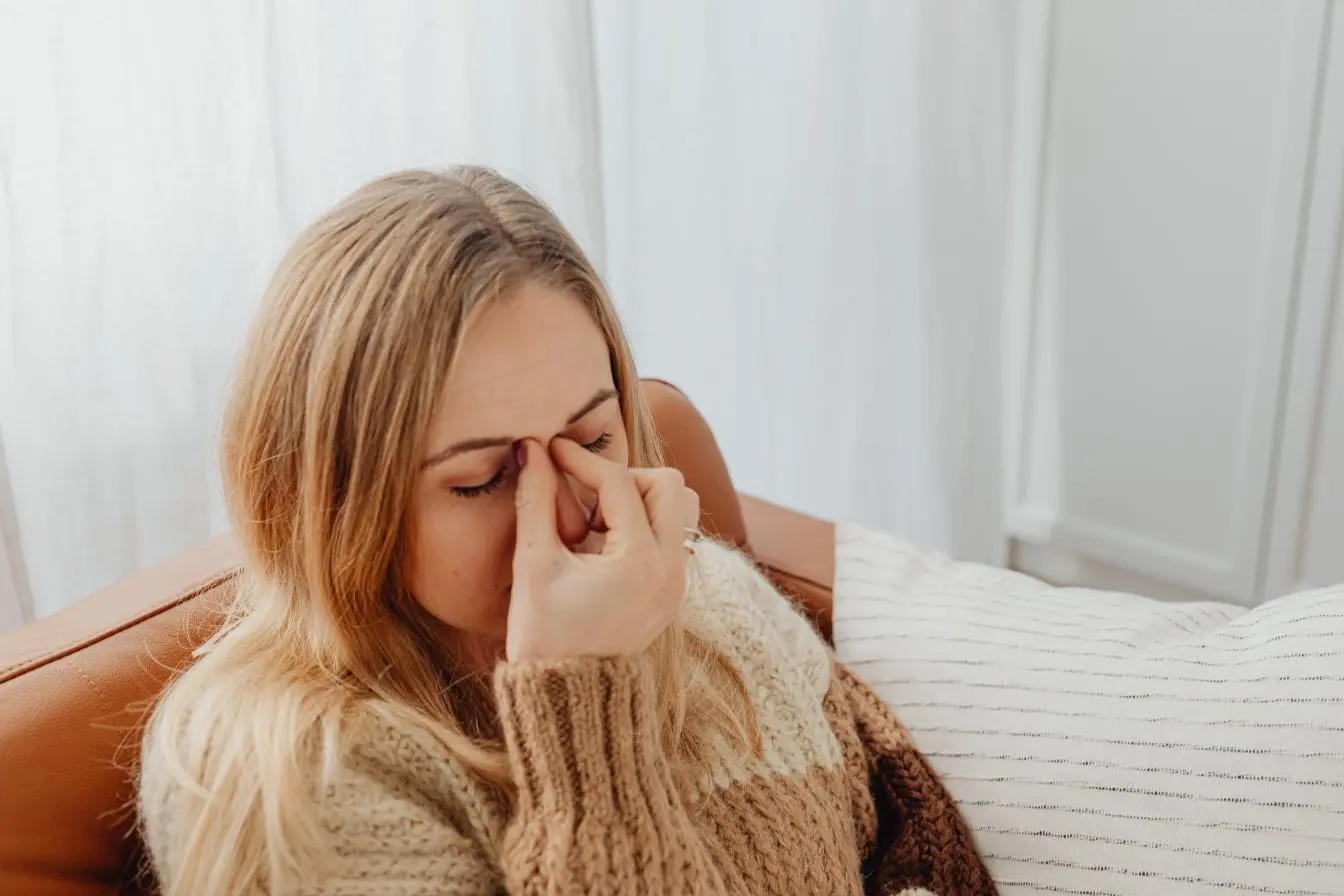 névralgie faciale Personne assise sur fauteuil en tricot beige, yeux fermés et doigts sur l’arête du nez, image qui illustre la douleur liée à une névralgie faciale.