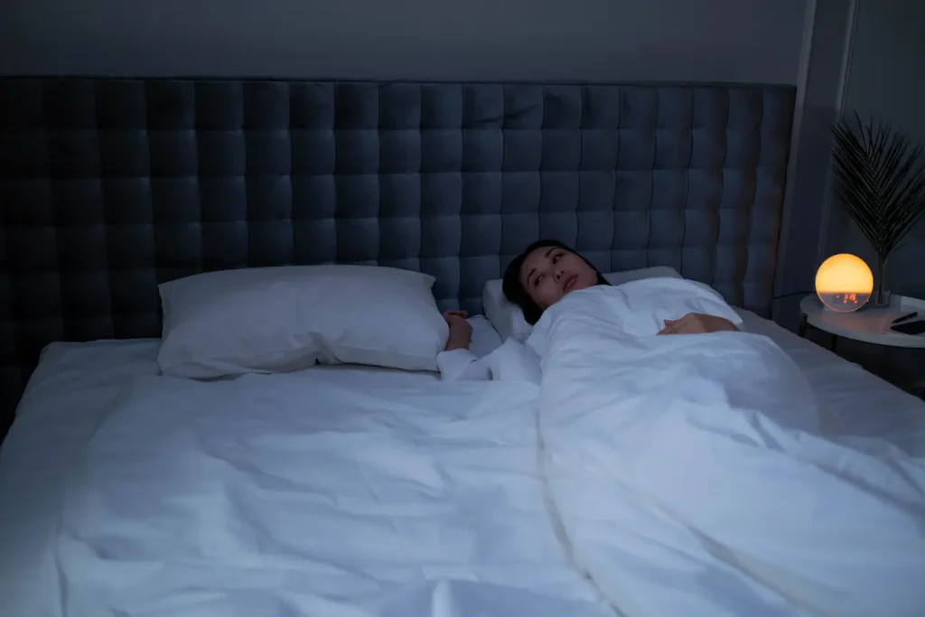 Femme allongée sous couette blanche dans chambre sombre, image qui illustre les sueurs nocturnes chez la femme et leurs causes.