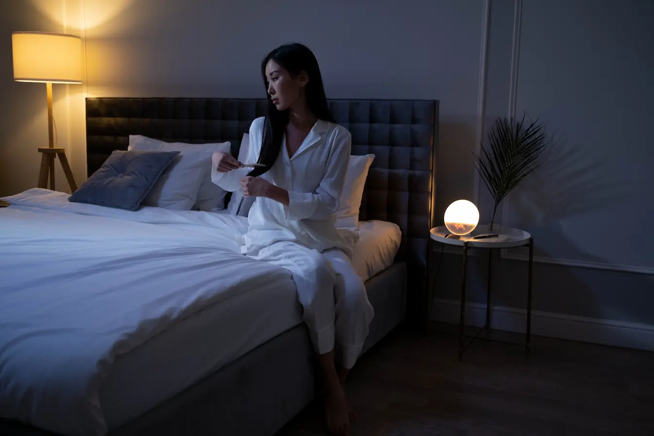 Femme en pyjama Femme en pyjama blanc assise au bord du lit dans chambre tamisée, image qui illustre les sueurs nocturnes chez la femme et leurs causes.