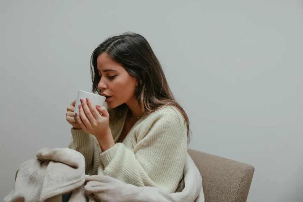 Personne assise enveloppée dans couverture beige tenant une tasse chaude, reflet des gestes pour soulager un mal de gorge.