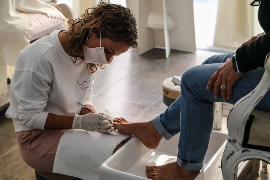 Personne en blouse blanche et masque soignant les pieds d’un client, image qui illustre le soin d’une mycose des ongles en salon.