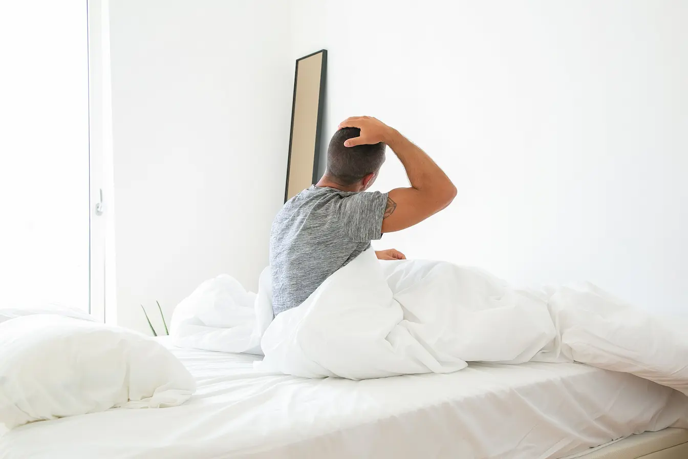 Personne assise sur lit Personne assise sur lit blanc, main posée derrière la tête, symbole des douleurs ressenties au réveil dans le dos.