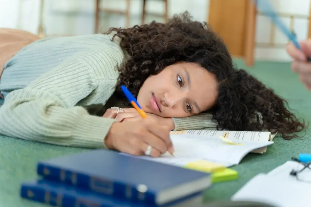 Une étudiante aux cheveux bouclés allongée sur le sol lutte contre la procrastination et le découragement en essayant d'étudier enfin.