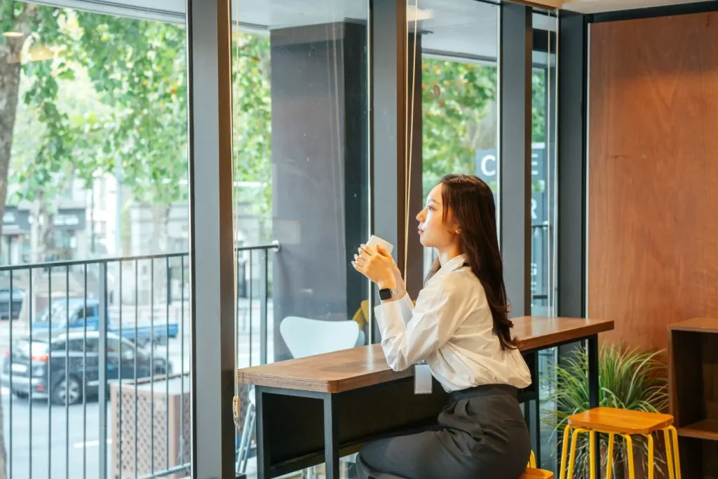 Une jeune femme seule assise à une table de café près d'une grande vitre, idéale pour illustrer le fait de gérer la solitude après une rupture en douceur.
