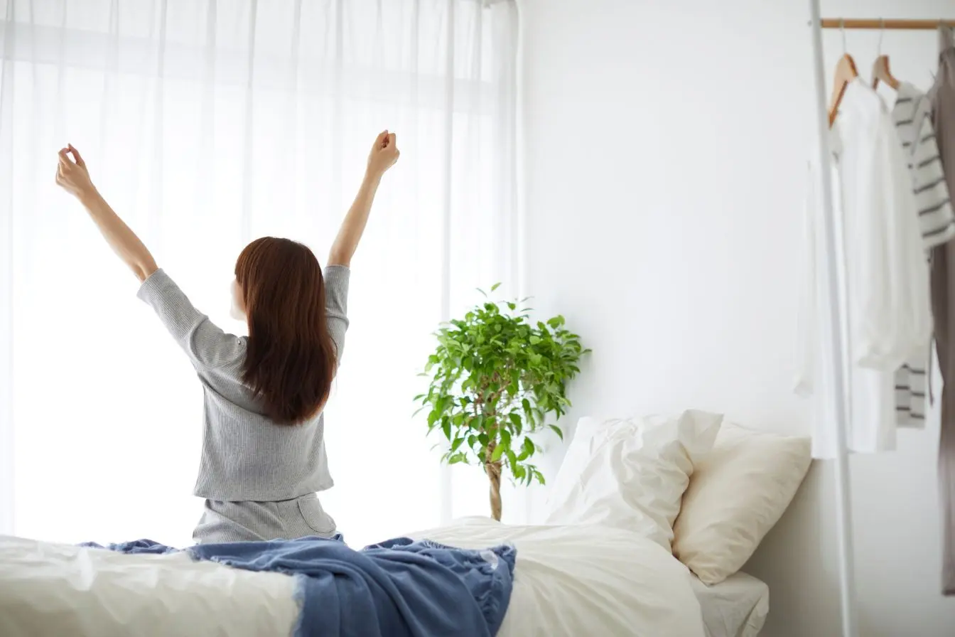 retrouver de l’énergie le matin Personne assise sur lit blanc levant les bras vers la fenêtre, symbole d’un réveil énergisant et naturel.